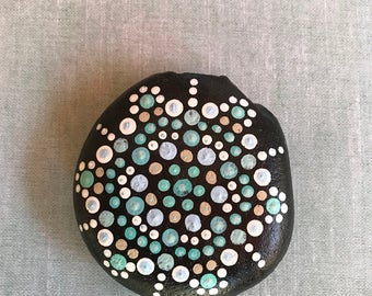 Dot painted mandala stone with blues, aquas, and white on a black background