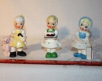 3 young lady figures by Ucagco Ceramics, Japan. One has a cake on a tray, one has a suitcase, and one has a teddy bear