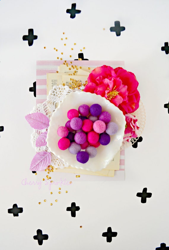 Felt balls in hues of blue, pink, and purple sit in a white bowl on top of doilies, book pages and flowers. The background is white with black crosses.  