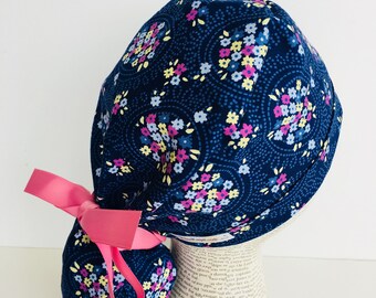 Ponytail Scrub Cap scrub hat featuring a dark blue material with flowers in blue purple and yellow with a matching pink ribbon