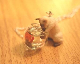 Mischievous Siamese Kitty cat is playing with goldfish in a fish bowl necklace