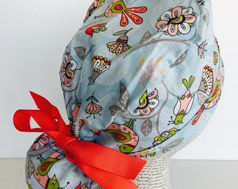 Ponytail Scrub Cap scrub hat featuring a light blue material with birds and flowers in red pink white and green with a matching red ribbon