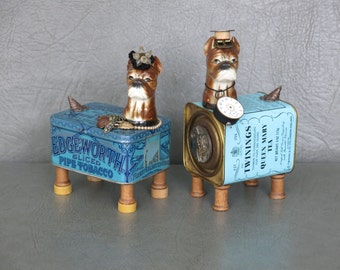 Pair of steampunk dog sculptures. Porcelain boxer heads affixed to vintage tins, spool feet, watch parts, gears, jewelry, found objects.
