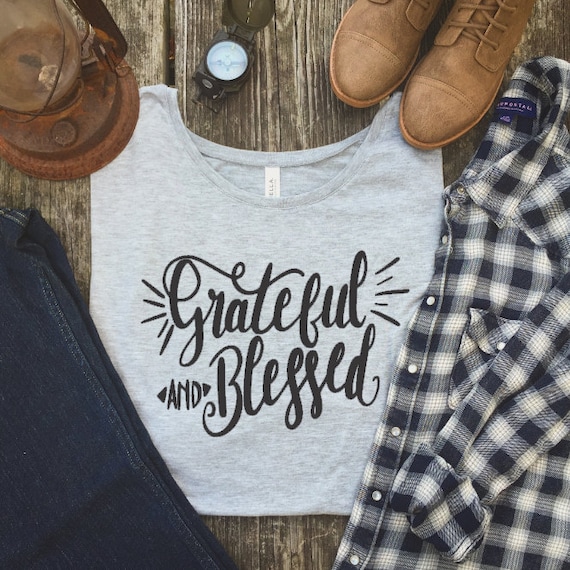 A screen printed t-shirt is styled with other clothing items. The gray t-shirt in the center says "grateful and blessed" in black lettering. Around it is a pair of old boots, a black and white plaid shirt, a pair of jeans, a vintage lantern, and an old compass.