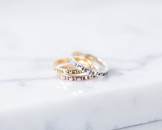 An image of three small rings with map coordinates are stacked on a white marble countertop. The rings are silver, gold, and brass colored.