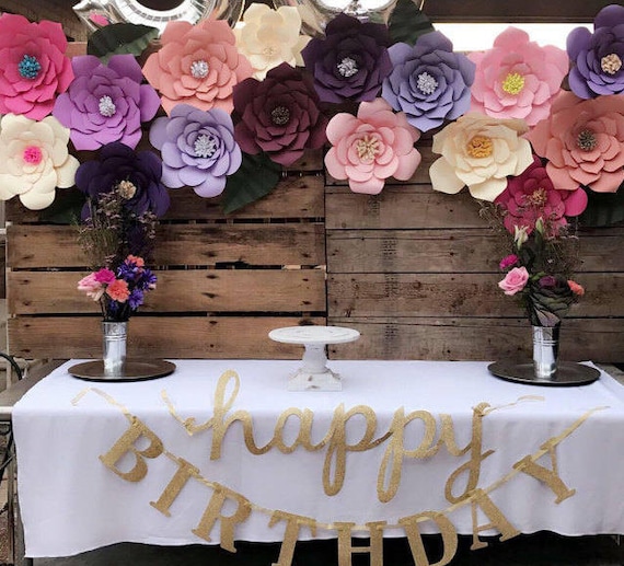 Giant paper flower are the backdrop to the party table set with a white tablecloth, with flowers and a large banner in gold glitter that reads "Happy Birthday." These party décor ideas can be found on Catching Colorflies.