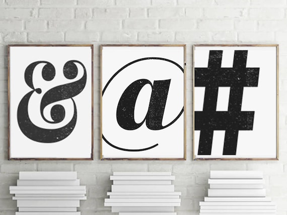 Three black and white framed wall art prints display an ampersand, at symbol, and pound sign, hung up on a white brick wall.
