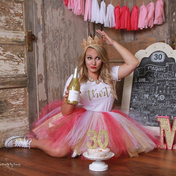 A picture from Raven's boutique shows a woman sitting on the floor in a pink tulle skirt and a white shirt with the words "thirty" in gold lettering. Paper tassels, a gold topped cake and a golden champagne bottle are in frame. 